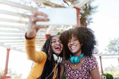 Smiling black woman with braids hugging cheerful african american female friend with curly hair and taking selfie together while standing on promenade in summer