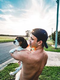 Shirtless man carrying dog while standing against sky