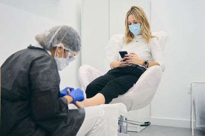 Female podologist in uniform doing pedicure for woman sitting in medical chair in beauty clinic