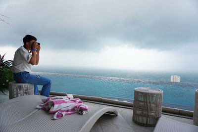 Side view of man photographing while sitting on seat against sea and sky