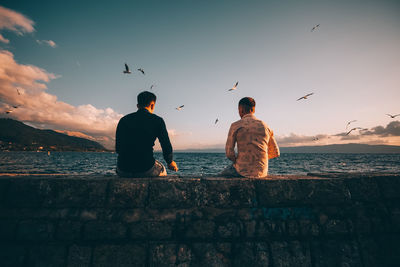 Men standing on retaining wall by sea against sky during sunset