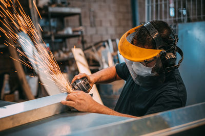 An adult man is seen working cutting a piece of metal in an industrial workshop, sparks are seen