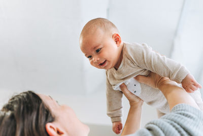 Young mother having fun with cute baby boy on hands in bright room, love emotion