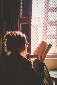 Woman reading book while sitting on bed by window