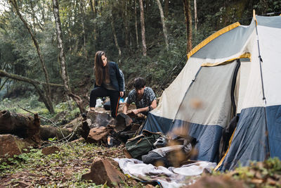 Couple of hikers making bonfire near tent surrounded by trees growing in forest while exploring nature together