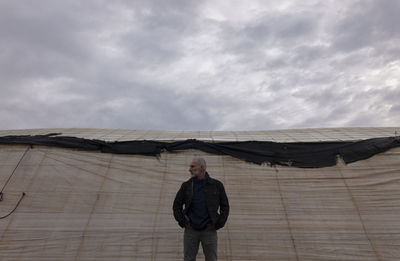 Adult man standing in front of plastic greenhouses against cloudy sky in almeria, spain
