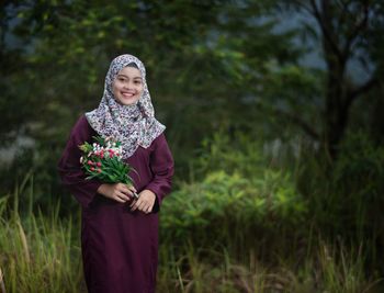 Portrait of happy woman in hijab holding flowers on field