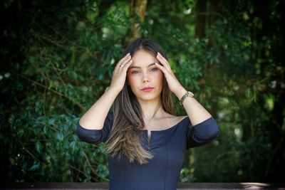 Portrait of beautiful young woman standing against plants seriously touching the hair