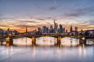 Illuminated bridge over river by buildings against sky during sunset