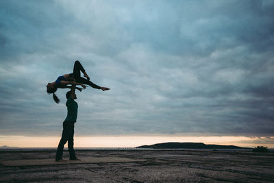 Full length of man balancing woman on hand by sea against cloudy sky