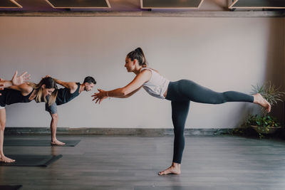 Slim barefoot female instructor in sportswear standing in warrior pose three and controlling group of sporty people concentrating and doing same exercise standing on sports mats in modern workout room