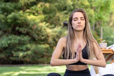 Front view of half cropped blonde lady meditating with hands together and garden view behind her