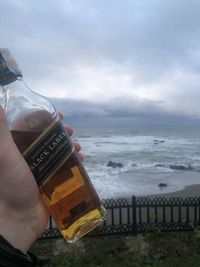 Person holding bottle by sea against sky