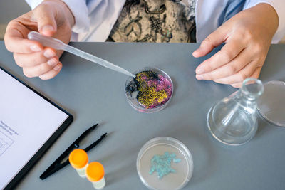 Female scientist adding drop of liquid from from pipette over petri dish with mix of glitters in lab
