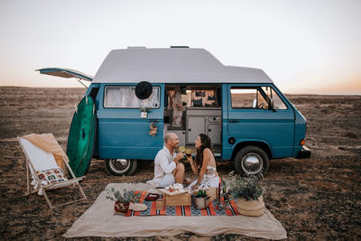 Side view of traveling couple sitting on blanket near van in savanna and having picnic while enjoying summer vacation and looking at each other