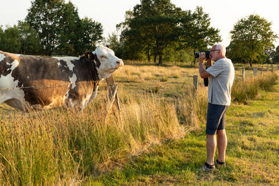 Side view of man photographing cow on grass against sky