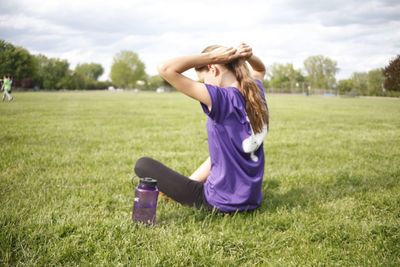 Girl tying hair while sitting on grassy field against sky