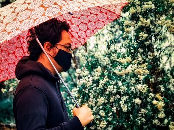 Midsection of man holding red umbrella against white flowering plants.