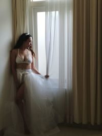 Seductive young woman wearing white dress looking through window at home