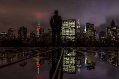 Rear view of man standing on glass floor against illuminated buildings