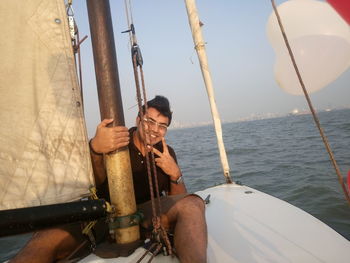 Portrait of young man gesturing peace sign while sitting in sailboat over sea
