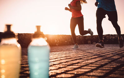 Low section of athletes running on footpath against sky at sunset