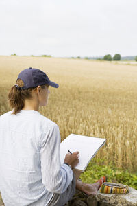 Woman drawing on paper while sitting in front of farm against clear sky