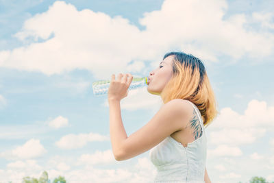 Young woman drinking water from bottle against cloudy sky