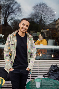 Portrait of smiling fashionable man standing with hands in pockets against cafe window