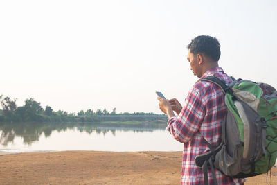 Side view of young man using phone by lake against sky