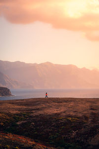 Remote side view of unrecognizable female explorer walking along rocky coast near sea on background of sunset sky and highlands on gran canaria