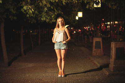 Portrait of young woman standing on footpath at night