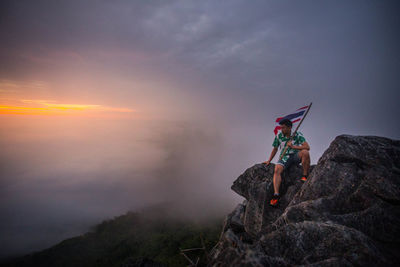 Man with flag sitting on rock during sunset