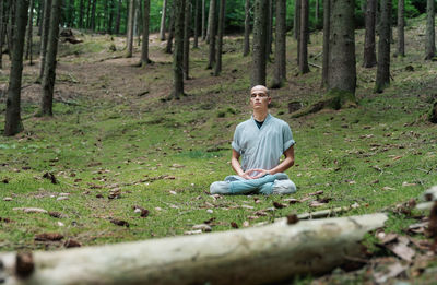 Bald man in traditional clothes sitting on rock in lotus pose and meditating during kung fu training in forest