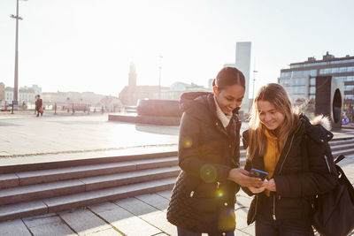 Smiling teenage friends looking at smart phone while standing in city during sunny day