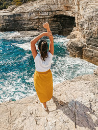 Young woman in yellow skirt standing on cliff above sea.