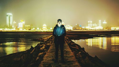 Portrait of young man standing amidst sea on groyne in city at night