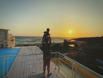 Rear view of man standing on railing against sea during sunset