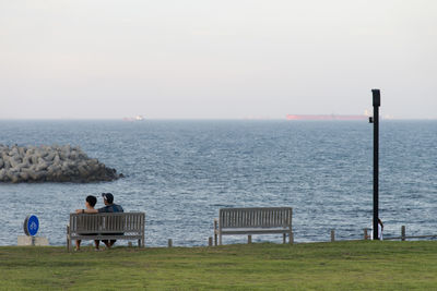 Rear view of couple sitting on bench at sea shore against clear sky