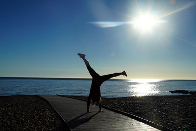 Full length of woman doing cartwheel on boardwalk at beach against sky during sunny day