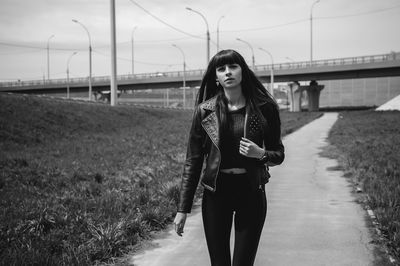 Portrait of fashionable young woman walking on footpath by field with bridge in background