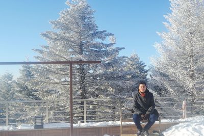 Smiling man sitting on bench against trees during winter