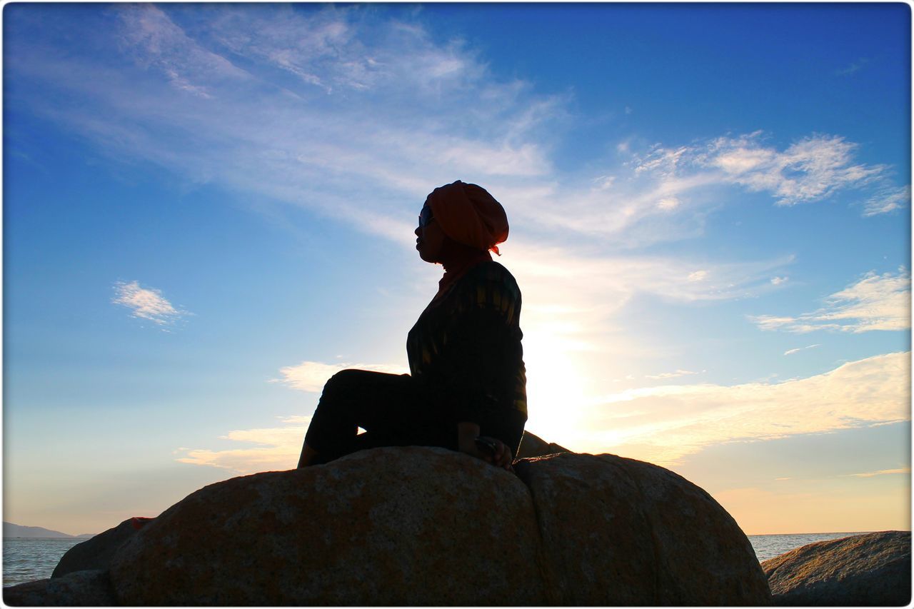 sky, lifestyles, standing, leisure activity, three quarter length, waist up, silhouette, cloud - sky, sunset, rock - object, cloud, rear view, casual clothing, sitting, looking at view, nature, low angle view, getting away from it all