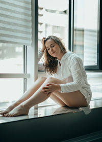 Portrait of young woman using phone while sitting on window