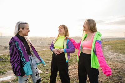 Group of teen hipster ladies in colorful sportive clothes laughing while chatting on empty meadow