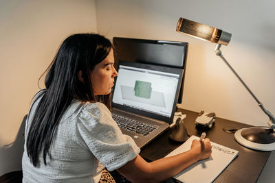 Rear view of young businesswoman sitting at desk, using laptop and 3d design software