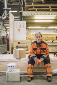 Full length portrait of smiling mature carpenter wearing reflective clothing while sitting in warehouse