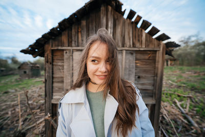 Beautiful girl with long hair in a grey trench coat next to an old wooden house outdoors in spring