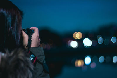 Portrait of woman photographing illuminated camera against sky at night