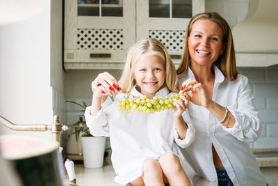 Happy blonde long hair mom and daughter having fun with grapes in the kitchen, healthy family 